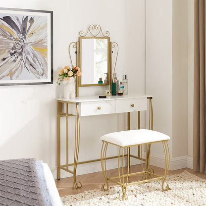 A white makeup dressing desk with gold-colored metal frame and legs, accompanied by a mirror and a padded stool. The room setting includes a bed and a nightstand with a lamp.