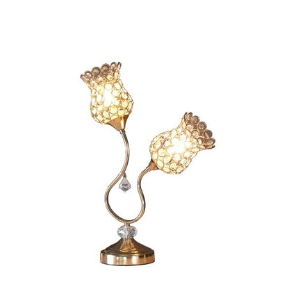 An ornate table lamp with a rose gold finish and a vintage floral metal design, featuring two flower horn motifs. The lamp is illuminated, casting a soft glow, and placed in a room setting with books and a picture frame in the background.