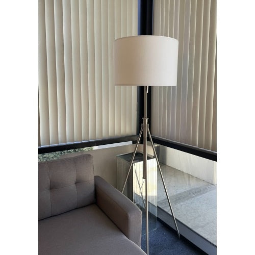 An adjustable tripod floor lamp with a white KD fabric shade, matte black stand, and brass-finish hardware, standing in a room with a gray couch and a window with vertical blinds.