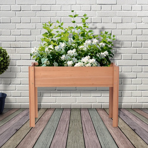 A wooden raised garden bed with legs containing white flowers, placed on a wooden surface with a brick wall in the background.