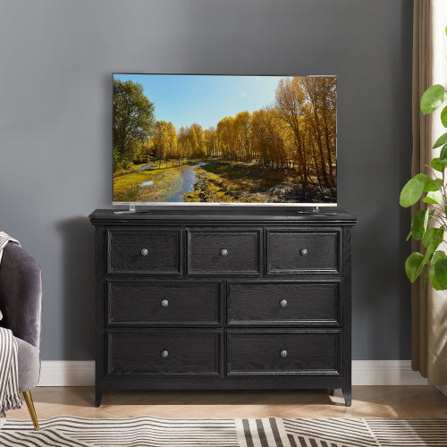 A black, handleless seven-drawer fiberboard vanity with an extended tabletop, situated in a well-lit room with a flat-screen TV on top displaying a nature scene, placed against a gray wall with a potted plant to the side.