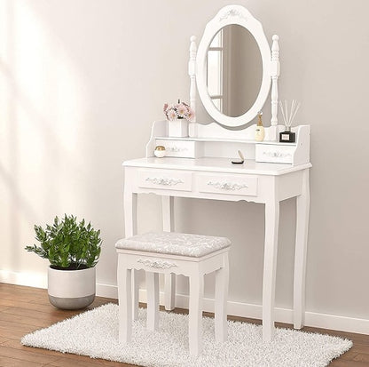 A white vanity table set with four drawers, an oval mirror, and a cushioned stool. The set is placed on a rug, next to a potted plant, on a hardwood floor against a white wall.