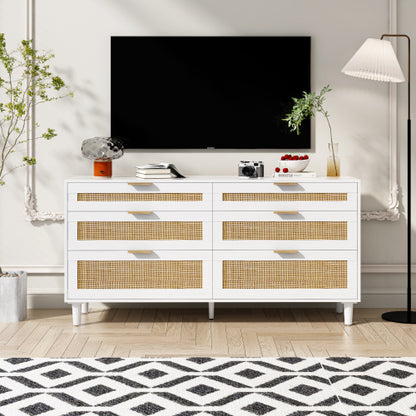 A white rattan dresser with six drawers featuring metal handles and wood legs, placed in a bedroom setting with a flat-screen TV on top, flanked by a plant and a lamp on either side.
