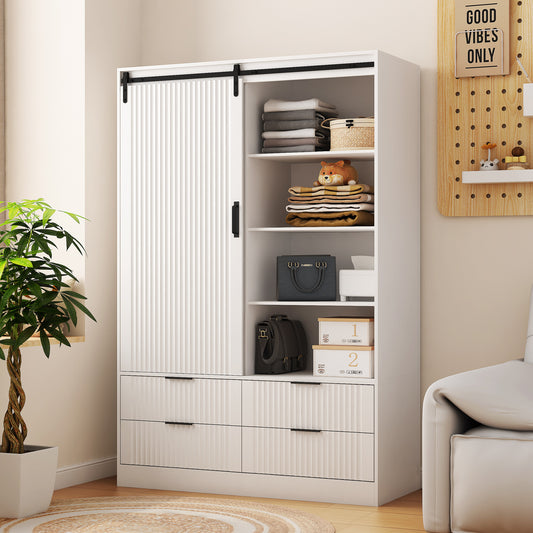 A white tall bedroom armoire wardrobe closet with sliding barn doors, drawers, and open shelves, placed in a room with a plant and a couch in the background.