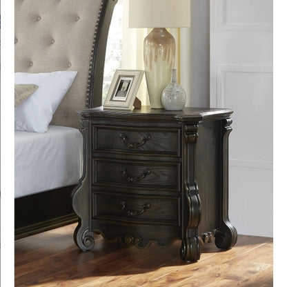 A traditional-style dark brown nightstand with intricate shaping and detailing, placed in a bedroom setting with a lamp and picture frame on top.
