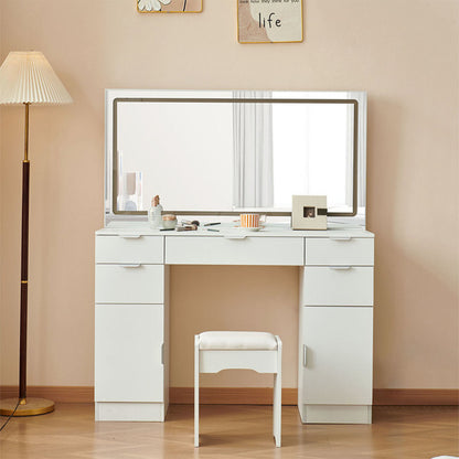 A modern white dressing table set with LED lighting, drawers, and a padded stool. The room it's placed in appears bright and well-lit, with a lamp in the background and a framed picture on the wall.