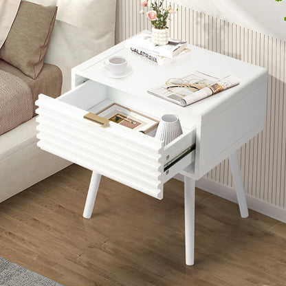 A white solid wood bedside table with a drawer and a slatted design, placed on a floor next to a couch, with a cup and a book on top of it.