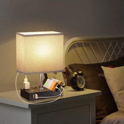 Table lamp on a nightstand next to a bed with pillows and an alarm clock.