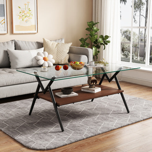 A rectangular coffee table with a transparent glass top and black metal legs placed on a grey area rug in a living room setting, with a sofa, cushions, and a decorative plant in the background.