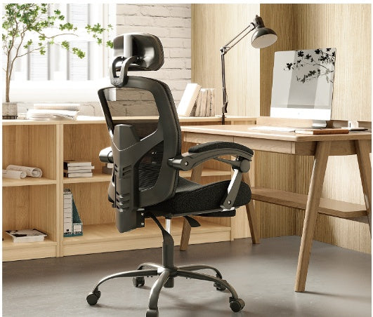 Black ergonomic swivel office chair with lumbar support pillow and padded armrests, featuring a high backrest and retractable footrest, positioned in a room with a wooden desk and bookshelf in the background.