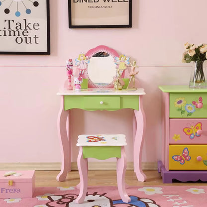 A colorful children's vanity set with a pink and green table, white mirror, and matching stool, placed in a room with various children's books and a decorated wall.