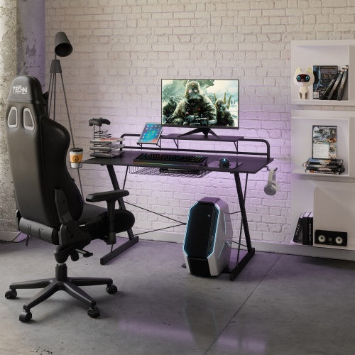 A black Techni Sport TS-200 Carbon Computer Gaming Desk with shelving, featuring a monitor stand, cup holder, and storage rack. The desk is pictured in a room with a gaming chair, a laptop, and various gaming accessories.