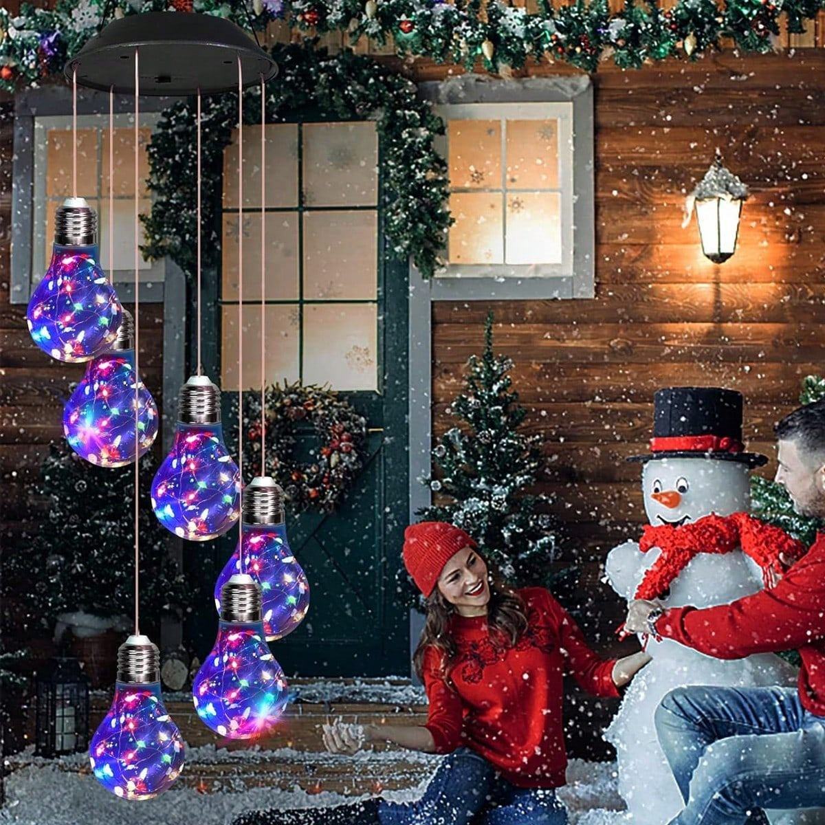 Two people in winter clothing sitting next to a snowman with colorful lighted bulb decorations in front of a wooden building.