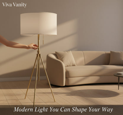 An adjustable tripod floor lamp with a white KD fabric shade, matte black stand, and brass-finish hardware, standing in a room with a gray couch and a window with vertical blinds.